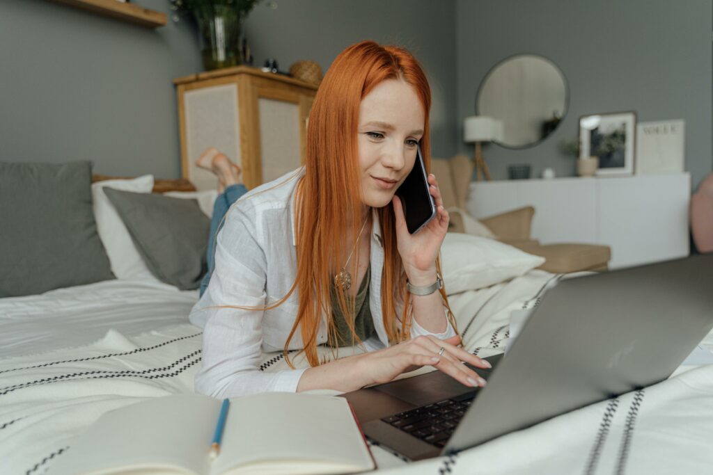 Red-haired woman multitasking in a cozy bedroom, using a laptop and phone for work.